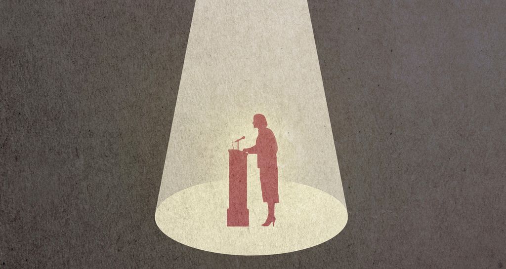Mot en mørk bakgrunn står en formelt kledd kvinne ved en talerstol. Hun står i spotlight fra en lyskaster over henne. Illustrasjon.