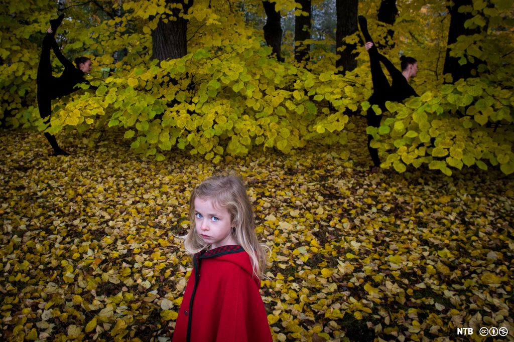 Lita jente med rød kåpe ser opp mot kamera. Hun er alvorlig. Gult høstløv i bakgrunnen. Foto. 