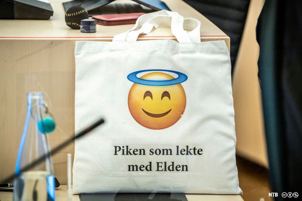 Et hvitt bærenett i tøy er stilt opp på en pult. Motivet er en emoji med glorie og teksten "Piken som lekte med Elden". Foto..